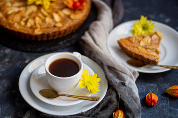 Charlotte apple pie in a round shape, decorated with fan-laden apple slices from the top, a piece of cake next to it, a cup of coffee, autumn leaves, all against a dark background. 
