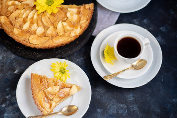 Charlotte apple pie in a round shape, decorated with fan-laden apple slices from the top, a piece of cake next to it, a cup of coffee, autumn leaves, all against a dark background. 