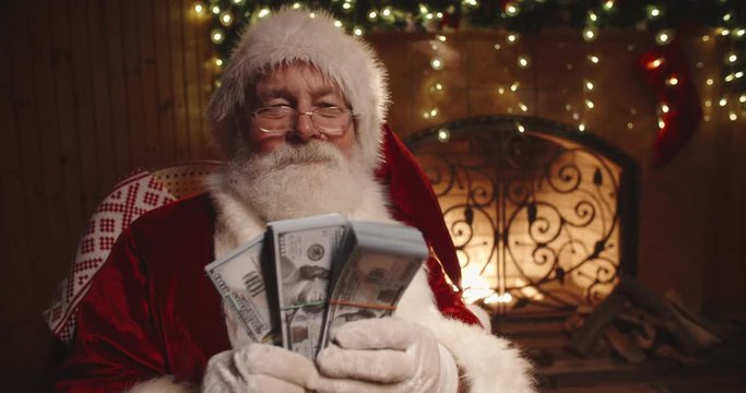 Authentic santa claus sitting in his rocker near fireplace and shaking packs of money in his hands - christmas spirit, holidays and celebrations concept 4k footage