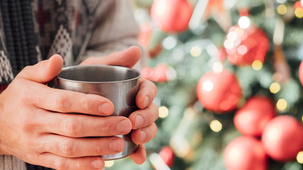 New year home celebration. Closeup of man hands holding hot drink in steel mug over blur decorated fir tree. Copy space.