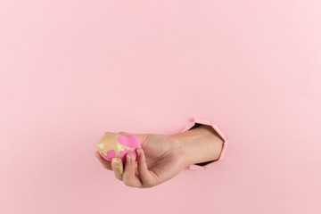Girl hand holds a beauty blender in foundation, concealer from a hole in a pink background. Makeup artist concept, copy space.