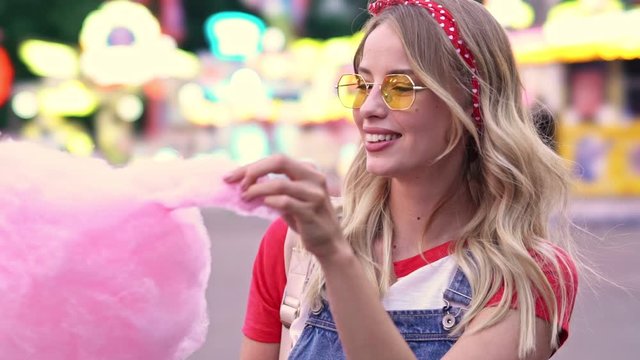 Close up view of attractive young blonde woman smiling and enjoying eating cotton candey while spending time at amusement park