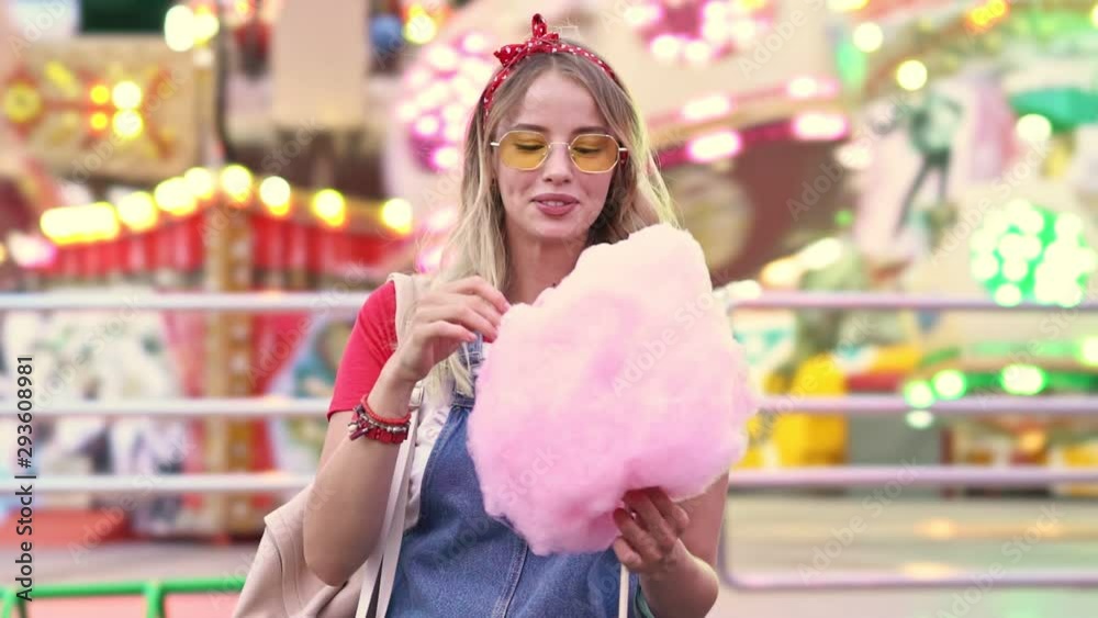 Wall mural Excited young blonde woman laughing and enjoying eating candy floss while having fun at amusement park