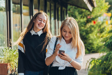 Fototapeta premium Happy moments of two stylish girls hugging and looking at the phone, discussing something on the street in the city. Close up portrait of cheerful joyful attarctive young girls best friends.