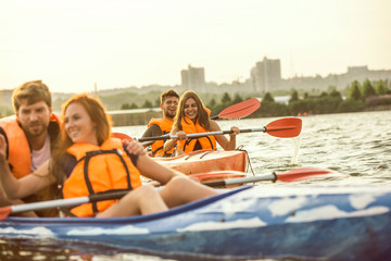 Obraz premium Happy young caucasian group of friends kayaking on river with sunset in the backgrounds. Having fun in leisure activity. Happy male and female model laughting on the kayak. Sport, relations concept.