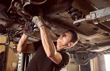 Repairman fixing car in garage. Experienced specialist car mechanic standing under lifted car during repair and maintenance process in repair station. Workshop for automobile checking up and repairing