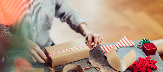Woman sitting by the table and wrapping Christmas presents with brown paper
