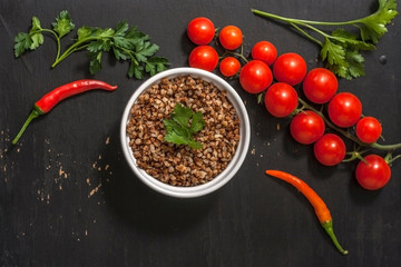 Cooked buckwheat porridge in a deep plate on a black wooden background with vegetables. Copy space. Top view