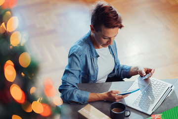 Woman sitting by the Christmas tree using smartphone and paying online with credit card