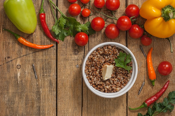 Cooked buckwheat porridge in a deep plate on a wooden background with vegetables. Top view