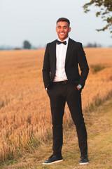 Young handsome guy in black suit with bow tie with his hands in his pockets and smiles, standing on a newly harvested arable land in the sunset