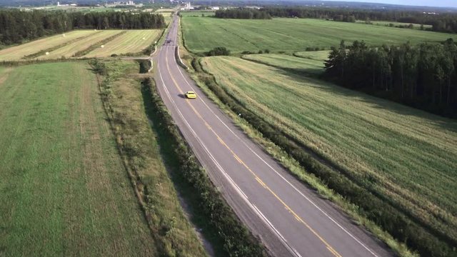 Aerial Chase Shot Of Yellow Muscle Car Entering Driveway On Remote Rural Road Traveling Through Beautiful Grass Fields On Sunny Summer Day