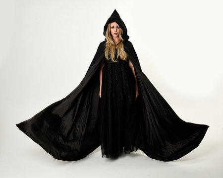 full length portrait of blonde girl wearing long black flowing cloak, standing pose  with  a white studio background.