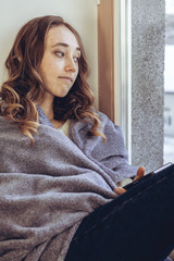 Sad woman in blanket sitting on window