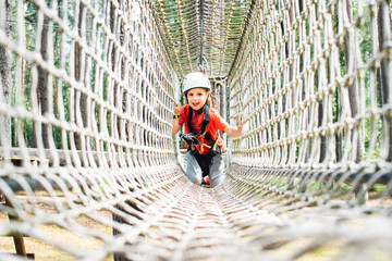 Little boy with climbing gear climbing rope trail between pine trees in adventure park. Boy enjoys climbing in the ropes course adventure. Rope park.