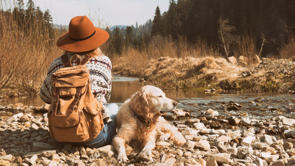 Traveling woman and wet golden retriever dog in nature. Stylish hipster in brown hat and wool sweater with vintage textile backpack near mountains river and forest.  Travel and wanderlust concept.