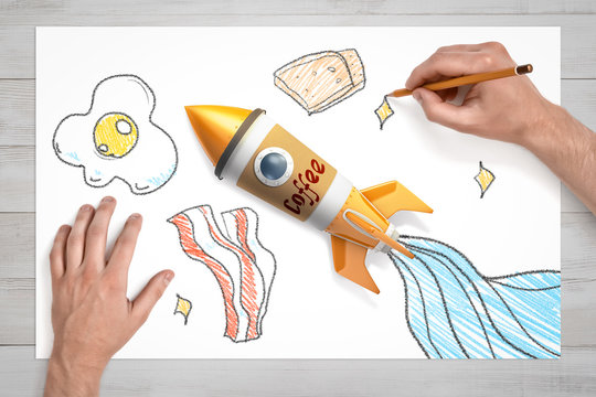 View from above of hands drawing bread, and eggs and bacon on white sheet of paper, with toy rocket made of coffee cup right in the centre.