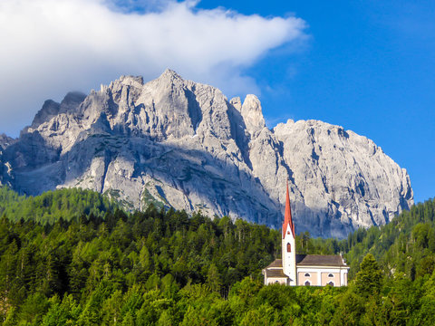 A church popping out of the forest, with a tall, sharp and rocky mountains behind. The church is build on a rock, making it taller than surrounding trees. Massive Alps in the back. Clear day.