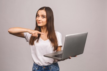 Surprised happy brunette woman in casual showing blank laptop computer screen and pointing on it while looking at the camera isolated over grey background.