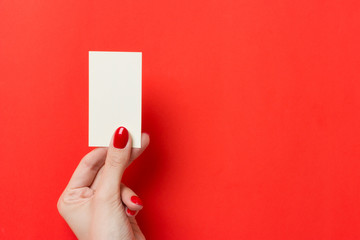 Female hands with red manicure holds a white blank business card on a red background.