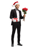 Young man in a suit and bow tie wearing a santa claus hat and holding roses and gift box