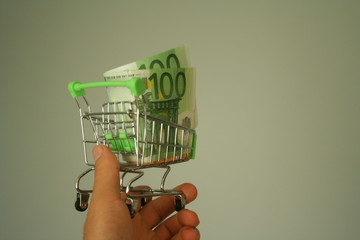 A mini dummy shopping cart in human hand with paper euro banknotes as a concept for online...