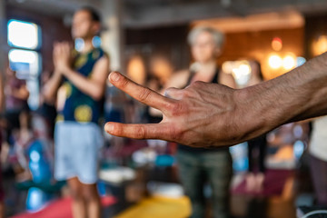 Fototapeta na wymiar Diverse group of people in yoga class. A close-up view on the hand of a young man giving the v symbol at the front of a workshop dedicated to sun salutations. Blurred practitioners are seen behind.