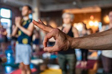 Fototapeta na wymiar Diverse group of people in yoga class. A motivational gym instructor is seen giving the ok hand gesture inside a gym during a yogic lesson as people are seen in standing pose in the background.