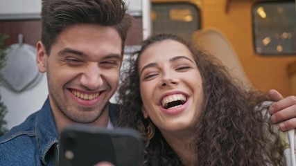 Close up view of happy beautiful young lovely couple hugging and taking selfie photo on smartphone while sitting near the trailer in the park - Powered by Adobe