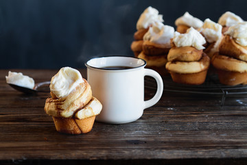 Fresh made homemade tall cinnamon roll with cream cheese icing. Hot steaming cup of coffee in the background with more buns cooling on a bakers rack over a rustic table.