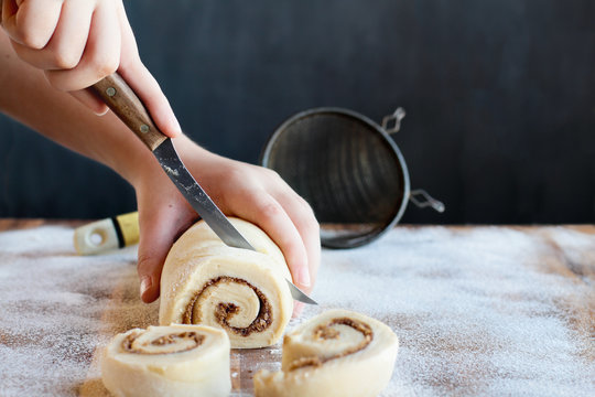 Woman's hands cutting homemade cinnamon roll dough over a floured surface with duster in background. Selective focus with blurred foreground and background. 