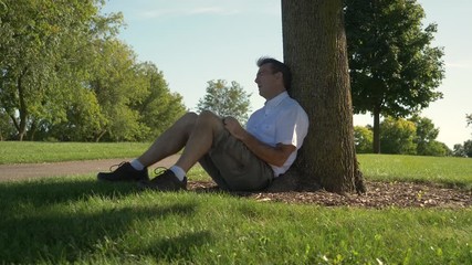 Man sleeping, sound asleep sitting at the base of a tree outdoors in a park on a warm sunny summer day. - Powered by Adobe