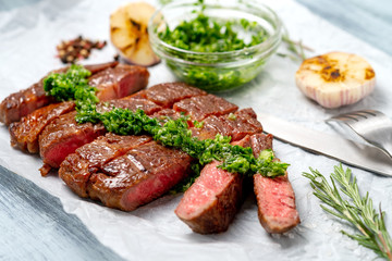 Sliced rib eye beef steak with chimichurri sauce and spices on white paper. Striploin cooked on the grill, top view