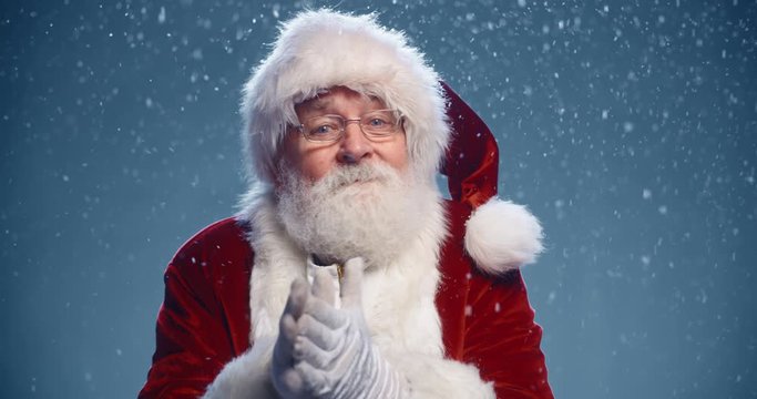 Senior man in santa clause outfit looking at camera, smiling and rubbing his hands, isolated on blue snowy background - christmas spirit concept close up 4k footage