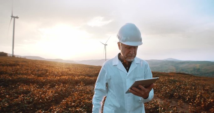 Mature caucasian engineer in hardhat and suit using tablet computer to check wind turbine system working at sunset on ecological station.