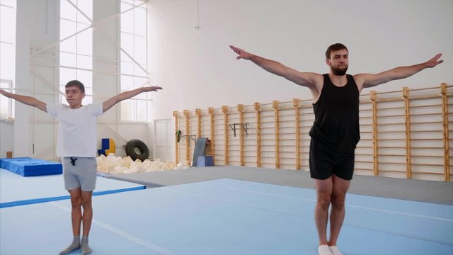 A cartwheel and a tripple back flip are made by a male sportman and a teen boy synchronously on the blue mats at sport centre, steadicam shot, back view.
