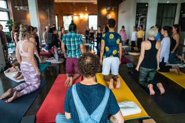 Diverse group of people in yoga class. A young man with short curly hair and a bald spot is seen sitting on a gym mat at the back of a room as people kneel in a circle and practice 108 sun salutes.