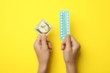Woman holding condom and birth control pills on yellow background, top view. Safe sex