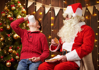 Santa Claus and child boy drinking tea, eating cookies, talking and sitting indoor near decorated xmas tree with lights - Merry Christmas and Happy Holidays!