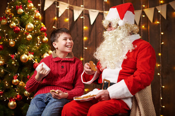 Santa Claus and child boy drinking tea, eating cookies, talking and sitting indoor near decorated xmas tree with lights - Merry Christmas and Happy Holidays!