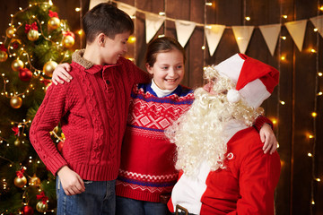 Santa Claus and child boy and girl posing together indoor near decorated xmas tree with lights, they talking, smiling and accepting gifts - Merry Christmas and Happy Holidays!