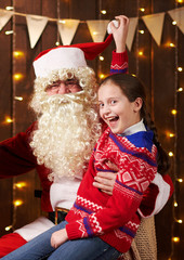 Santa Claus and child girl posing together indoor near decorated xmas tree with lights, they talking and smiling - Merry Christmas and Happy Holidays!