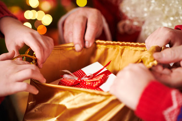 Santa Claus opens a bag and gives gifts to children, sitting indoor near decorated xmas tree with lights - Merry Christmas and Happy Holidays!