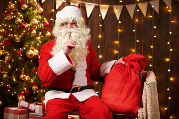 Santa Claus posing with bag of gifts, sitting indoor near decorated xmas tree with lights - Merry Christmas and Happy Holidays!