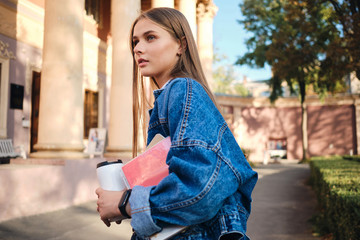 Side view of casual student girl with cup to go and textbook thoughtfully looking away outdoor