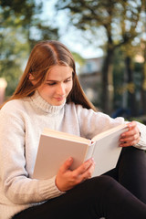Obraz premium Young pretty student girl in cozy sweater dreamily reading book while studying outdoor