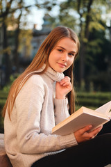 Obraz premium Young pretty smiling student girl in cozy sweater sensually looking in camera while studying with book outdoor