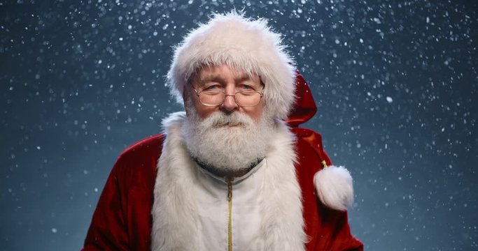 Santa Clause looking at camera when suddenly getting surprised and throwing his arms wide, isolated on snowy blue background - christmas spirit concept close up 4k footage