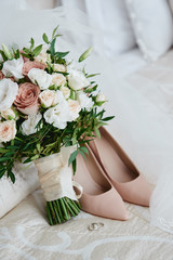 Bridal bouquet of pink, white roses and greenery with satin ribbon, beige women shoes, veil and two golden wedding rings on bed, copy space. Wedding concept