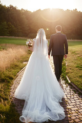 Full length body portrait of young bride and groom walking on green grass of golf course at sunset,...